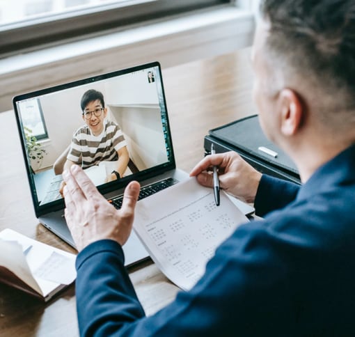 A man in a meeting video call with another man, with his notepad and pen in hand